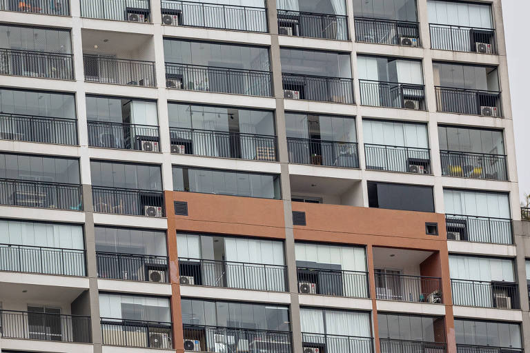 A imagem mostra a fachada de um edifício residencial com várias janelas de vidro. Quase todas as janelas possuem unidades de ar-condicionado instaladas. Há um destaque em uma parte da fachada, onde uma seção é pintada de laranja, contrastando com o restante da estrutura que é predominantemente branca e de vidro.