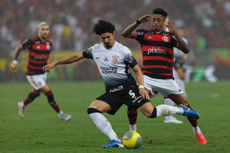 Jogador de futebol com uniforme branco e preto domina a bola com o pé direito， enquanto um adversário com uniforme vermelho e preto tenta desarmá-lo. Outro jogador com o mesmo uniforme do adversário observa ao fundo， em um estádio lotado com torcedores.







