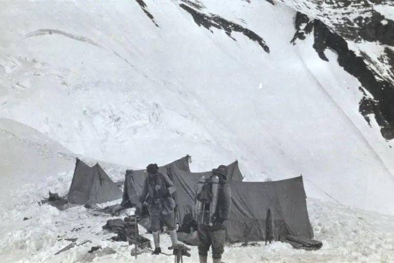 Dois jovens em meio à neve em foto em preto e branco. Atrás deles, duas barracas de tecido