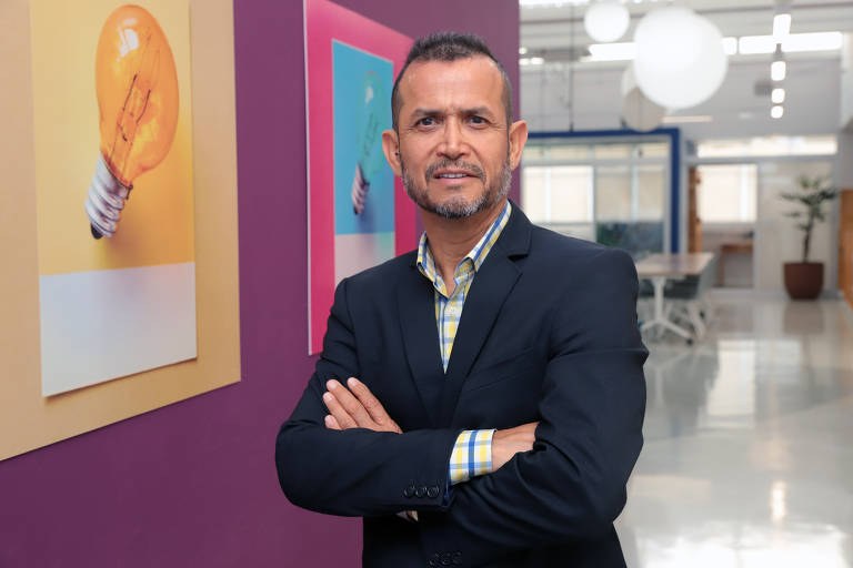 Um homem branco de terno preto e camisa de padrão xadrez, com braços cruzados, posa em um ambiente de escritório. Ao fundo, há duas ilustrações de lâmpadas em cores amarelo e azul, e o espaço é iluminado com luz natural, apresentando mesas e plantas.