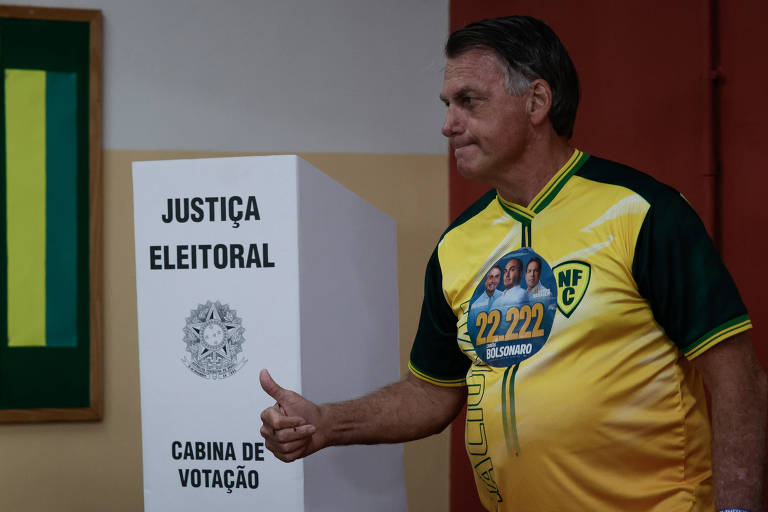 Um homem está ao lado de uma cabine de votação marcada com a inscrição 039;JUSTIÇA ELEITORAL039;. Ele usa uma camiseta amarela e verde， típica da seleção brasileira de futebol， e faz um gesto de positivo com a mão.