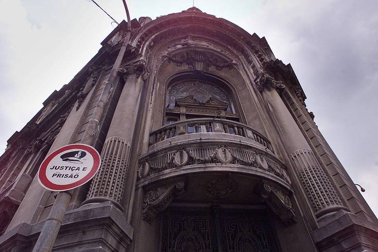 A imagem mostra a fachada de um edifício histórico， vista de baixo para cima. O prédio apresenta detalhes arquitetônicos elaborados， com uma grande entrada central e varandas decorativas. À esquerda， há um poste de sinalização com um aviso que diz 039;JUSTIÇA E PRISÃO039;. O céu está nublado， contribuindo para uma atmosfera sombria.
