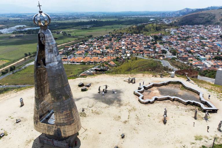 A imagem mostra uma grande estátua de Nossa Senhora Aparecida, localizada em uma colina. A estátua é metálica e possui uma coroa com uma cruz no topo. Ao fundo, há uma vista panorâmica de uma cidade com várias casas e áreas verdes. O céu está claro e há algumas nuvens. No chão, há uma estrutura em forma de lago, além de outras figuras e esculturas ao redor da estátua.