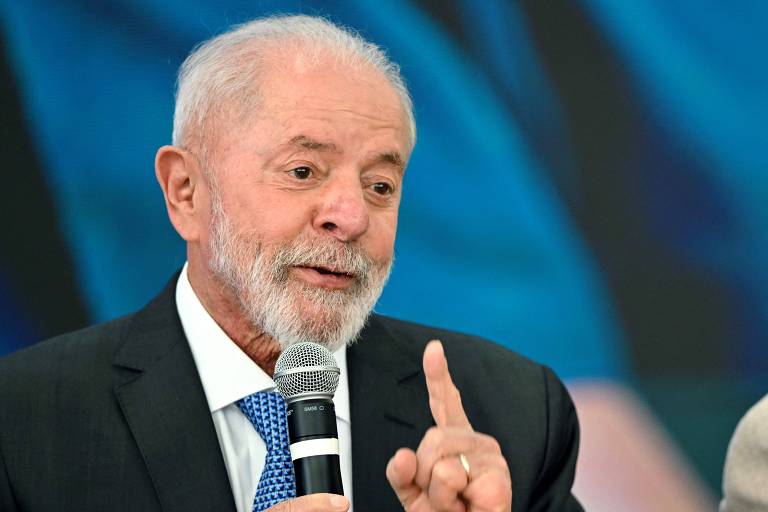 Um homem com cabelo grisalho e barba, vestindo um terno escuro e uma camisa branca, está falando ao microfone. Ele gesticula com a mão levantada, indicando um ponto importante. Ao fundo, há uma tela com um padrão azul.