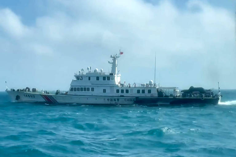 A imagem mostra um navio de grande porte navegando em águas azuis. O navio é predominantemente branco com detalhes em azul e vermelho. Há várias pessoas visíveis no convés, e o céu está parcialmente nublado.