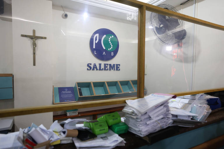 A imagem mostra o interior de um escritório com uma mesa desorganizada cheia de papéis e documentos. Ao fundo, há uma parede com um logotipo que diz 'PCS SALEME' e uma cruz na parte superior esquerda. O ambiente parece ser um espaço de trabalho com prateleiras visíveis e uma janela de vidro.