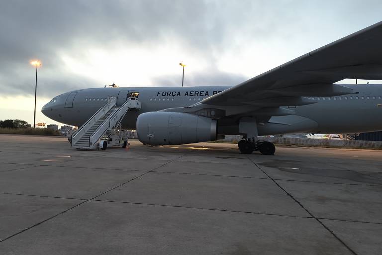 A imagem mostra um avião da Força Aérea estacionado em uma pista. O avião é de grande porte, com uma escada de embarque posicionada ao lado. O céu está nublado e há luzes de um aeroporto ao fundo.