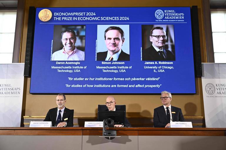  imagem mostra um painel de anúncio do Prêmio Nobel de Economia. No centro, há uma tela grande exibindo fotos de três pessoas.. Abaixo da tela, sentados a uma mesa, estão três homens de terno. O fundo da sala é branco com o logotipo do Prêmio Nobel visível.