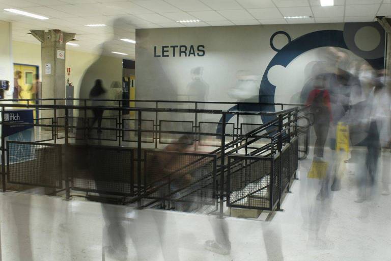 A imagem mostra um corredor da FFLCH, com a palavra 'Letras' escrita em letras grandes na parede. O ambiente é movimentado, com várias pessoas em movimento, criando um efeito de desfoque. Há uma escada com corrimão e uma área com grades. O piso é claro.