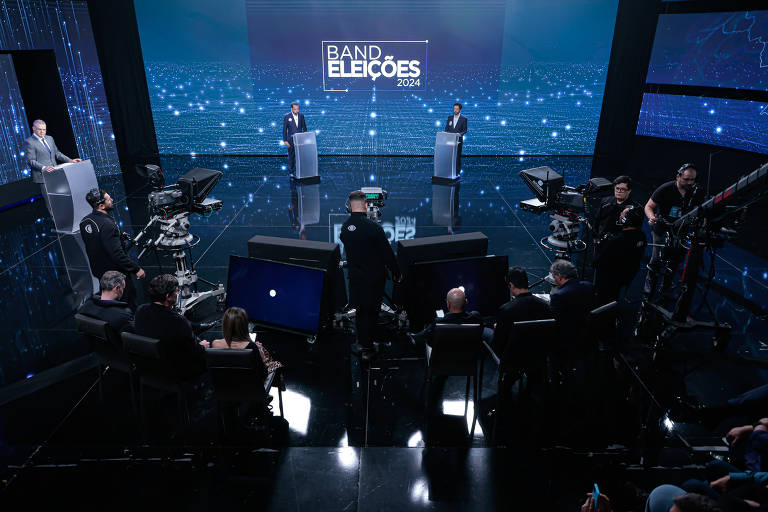 A imagem mostra um estúdio de televisão durante um debate eleitoral. Dois candidatos estão em pé atrás de púlpitos, enquanto câmeras e uma equipe técnica estão posicionadas em primeiro plano. O fundo é digital, com a palavra 'BAND' e 'ELEIÇÕES 2024' visíveis. A iluminação é focada nos candidatos, criando um ambiente de destaque.