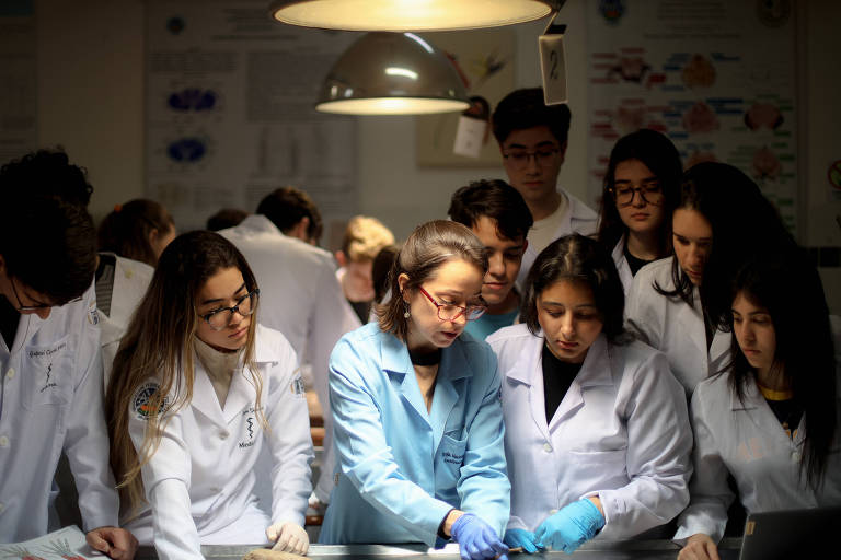 A imagem mostra um grupo de estudantes em um ambiente de aula prática, todos usando jalecos brancos. Eles estão reunidos em torno de uma mesa, observando e interagindo com um objeto que não é visível na imagem. A iluminação é suave, com uma lâmpada pendente acima deles. O grupo é composto por homens e mulheres, com expressões de concentração e interesse.