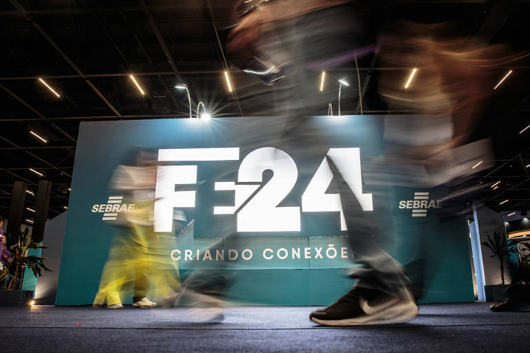 A imagem mostra um painel de fundo em um evento, com a inscrição 'FE24' em destaque, acompanhada da frase 'CRIANDO CONEXÕES'. Pessoas em movimento passam em frente ao painel, criando um efeito de desfoque. O ambiente parece ser um espaço de convenções ou feira.