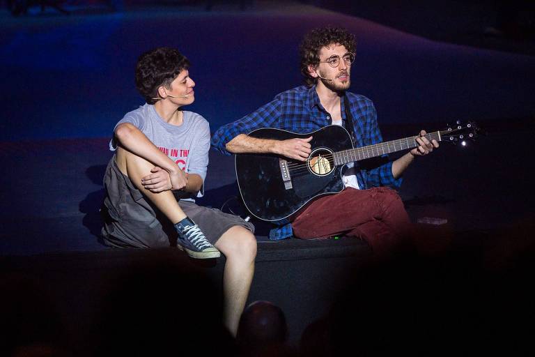 A imagem mostra duas pessoas em um palco durante uma apresentação musical. À esquerda, uma pessoa sentada com as pernas cruzadas, vestindo uma camiseta cinza e calças curtas, olhando para a direita. À direita, outra pessoa está tocando um violão preto, vestindo uma camisa xadrez azul e calças vermelhas. Ambas as pessoas têm microfones de lapela e estão em um ambiente com iluminação suave.