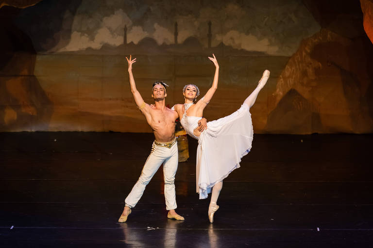 Um par de dançarinos de ballet se apresenta em um palco. O dançarino masculino está sem camisa, vestindo calças brancas e uma faixa dourada na cabeça, enquanto a dançarina feminina usa um vestido branco e sapatilhas de ponta. Ela está em uma pose de dança, com uma perna levantada e um sorriso, enquanto ele a segura com uma das mãos e levanta a outra em uma pose elegante. O fundo do palco apresenta uma pintura de paisagem com montanhas e nuvens.