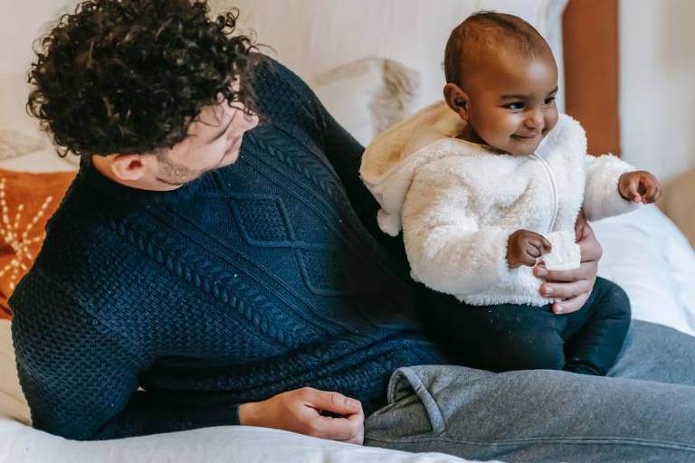 Homem branco com suéter azul escuro olha para bebê, de pele mais escura e jaqueta branca, que senta em seu colo