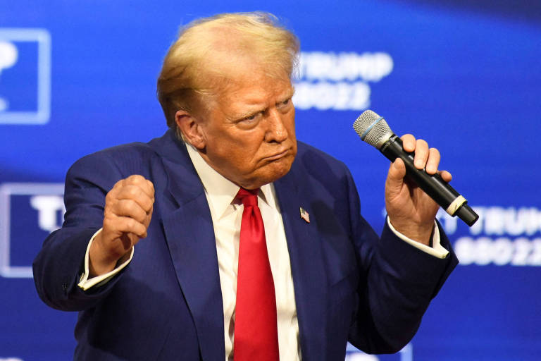 Trump, com cabelo loiro e terno azul, está segurando um microfone enquanto faz gestos com as mãos. Ele está em um evento com um fundo azul.