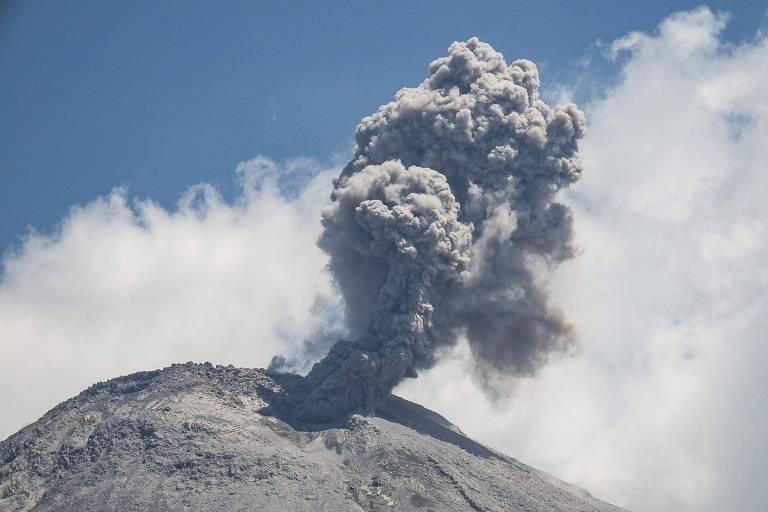 A imagem mostra um vulcão em erupção, com uma grande coluna de fumaça e cinzas sendo expelida para o céu. O vulcão está coberto por rochas e detritos, e o céu ao fundo é azul com algumas nuvens brancas.