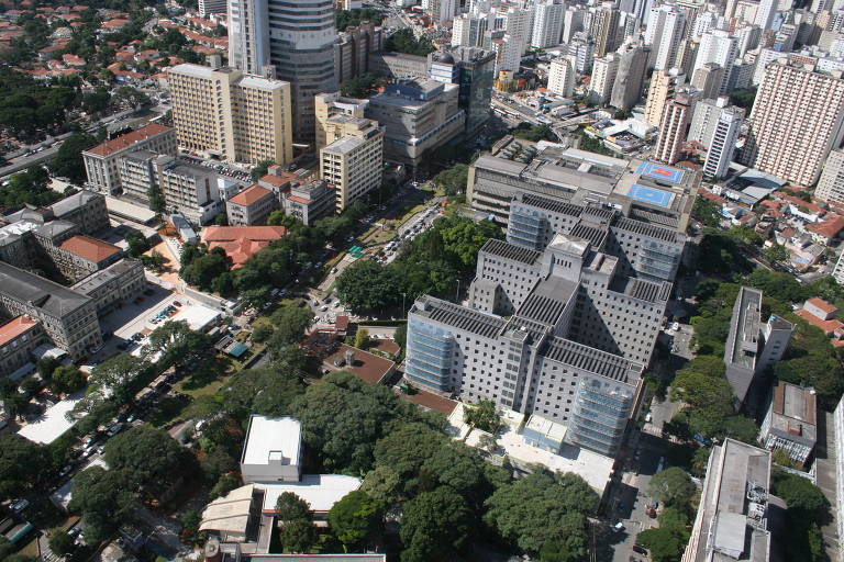 Imagem aérea de uma área urbana, mostrando uma combinação de edifícios altos e baixos, com áreas verdes intercaladas. Há uma variedade de estruturas arquitetônicas, incluindo prédios comerciais e residenciais, além de ruas e calçadas visíveis entre as construções.