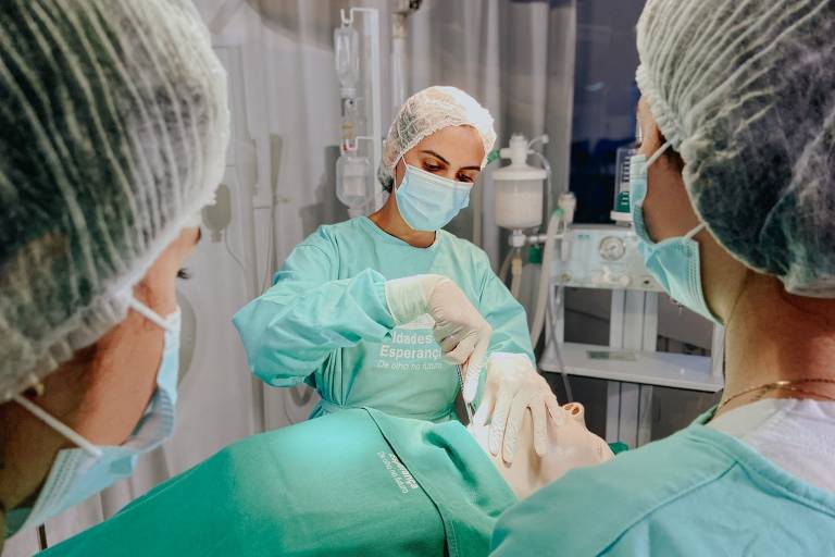 A imagem mostra uma equipe cirúrgica em um ambiente de operação. Um cirurgião， vestido com um avental verde e máscara， está realizando um procedimento em um paciente deitado na mesa de cirurgia. Dois outros profissionais， também com aventais verdes e máscaras， observam o procedimento. O ambiente é esterilizado e contém equipamentos médicos ao fundo.