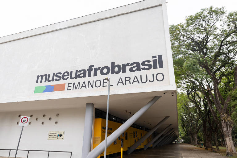 A imagem mostra a fachada do Museu Afro Brasil, com o nome 'museuafrobrasil' e 'EMANOEL ARAUJO' em letras grandes e coloridas. O edifício é moderno, com uma estrutura que se destaca por seus pilares. Ao fundo, há árvores que cercam o museu no parque Ibirapuera