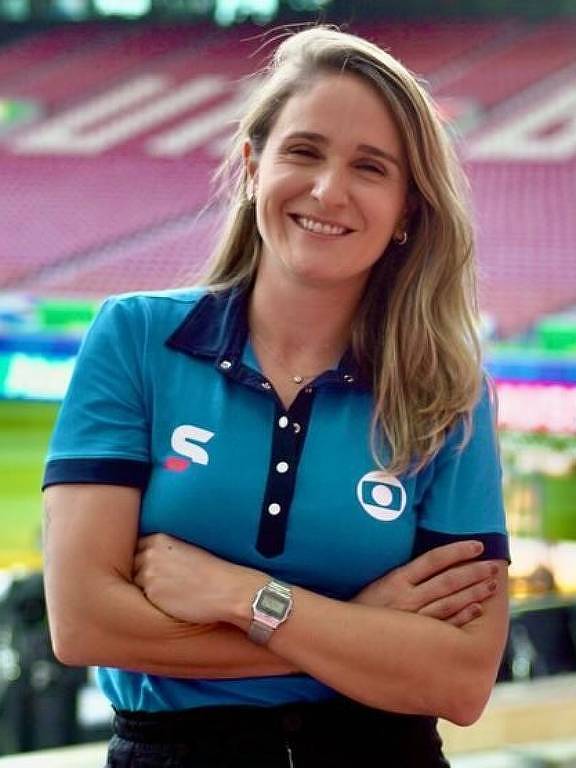 Uma mulher sorridente está em pé em um estádio de futebol, com os braços cruzados