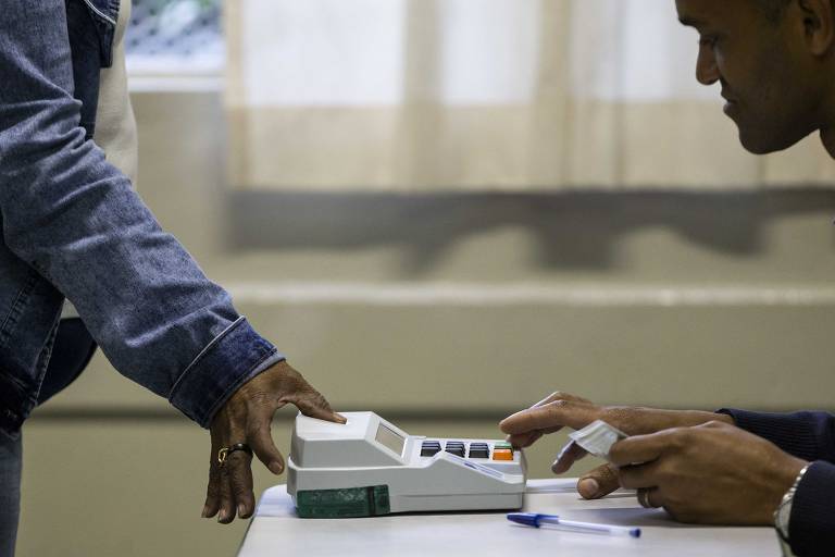 Foto mostra uma pessoa em pé de pele escura, de camisa ou jaqueta jeans, coloca a digital do dedão em máquina da Justiça Eleitoral branca com botões pretos. Do lado direito há uma outra pessoa também de pele escura, sentada. Estão em sala fechada, clara