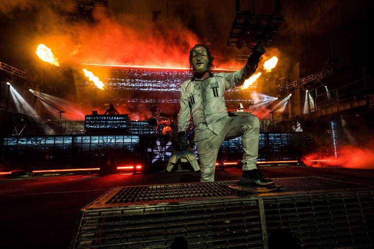 Um artista de música ao vivo está em um palco, vestindo um traje branco e uma máscara. Ele está em uma posição dinâmica, com uma mão levantada, enquanto chamas e luzes vermelhas iluminam o fundo. 