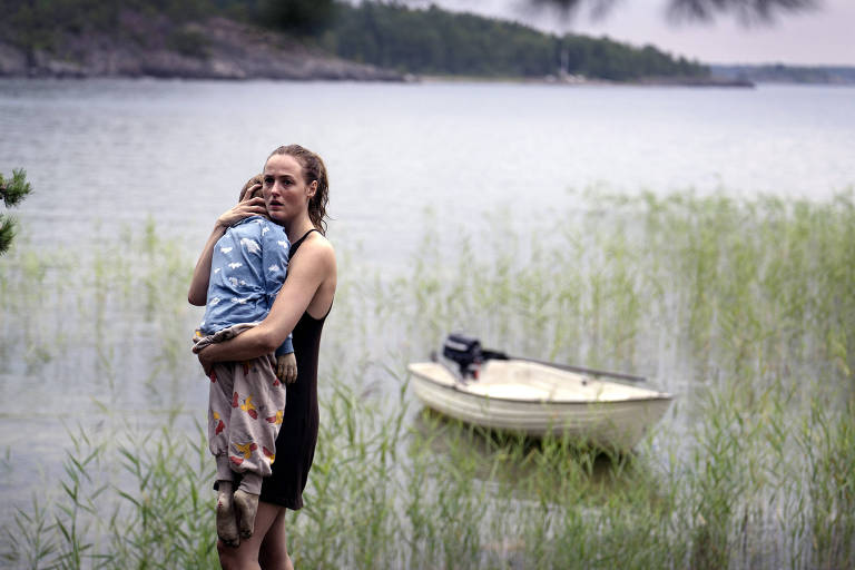 Mulher segura criança de camisa azul e calça na beira de um lago, ao lado direito dela, um pequeno barco