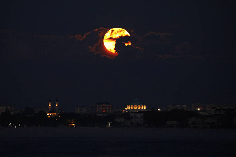 A imagem mostra uma lua cheia brilhante surgindo entre nuvens escuras no céu noturno. A lua tem uma coloração alaranjada e está parcialmente oculta por nuvens. Abaixo, é possível ver uma silhueta de uma cidade com edifícios iluminados, refletindo a luz da lua.