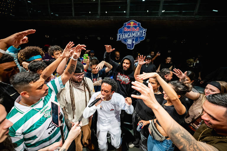 A imagem mostra um grupo de pessoas em um evento de dança, com várias pessoas levantando as mãos e se movendo em torno de um dançarino central. O dançarino está vestido de branco e parece estar no meio de uma performance. Ao fundo, há um logotipo da Red Bull