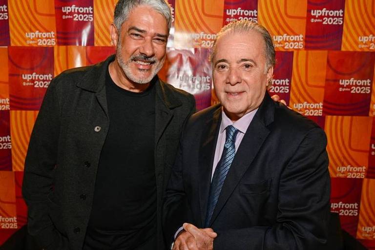 Duas pessoas posam para a foto em um evento chamado Upfront 2025. O fundo é composto por painéis coloridos com o logo do evento. A pessoa à esquerda tem cabelo grisalho e usa uma jaqueta escura, enquanto a pessoa à direita está vestida com um terno escuro e uma camisa clara. Ambas estão sorrindo e se cumprimentando.