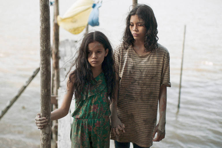 A imagem mostra duas meninas em pé à beira de um rio. A menina à esquerda usa um macacão verde com estampas florais e tem cabelo longo e solto. A menina à direita veste uma camiseta listrada e parece estar suja. Ambas estão segurando as mãos e têm expressões sérias. Ao fundo, é possível ver a água do rio e alguns postes de madeira.