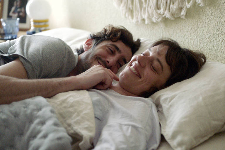 Um casal está deitado na cama, sorrindo um para o outro. O homem, com cabelo cacheado e barba, está deitado de lado, enquanto a mulher, com cabelo liso e curto, está deitada de costas. Ambos parecem felizes e relaxados, com lençóis claros e almofadas ao redor. Ao fundo, há uma parede texturizada e uma luminária.