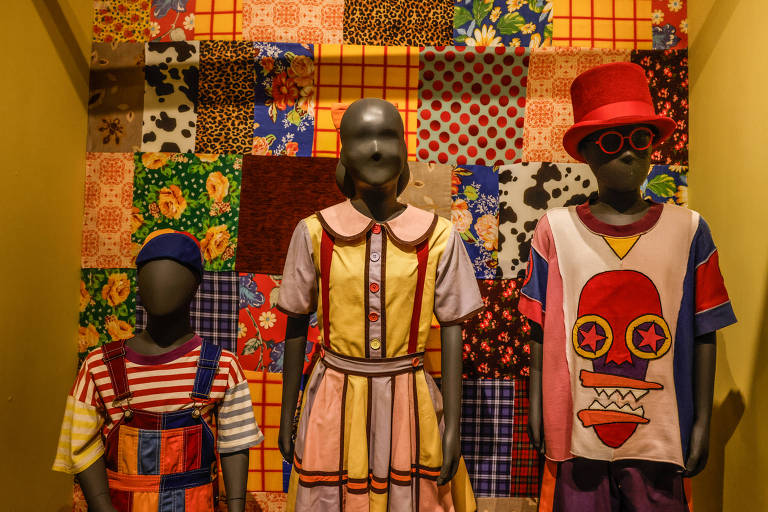 A imagem mostra três manequins em uma exposição de roupas. O manequim à esquerda está vestido com uma camiseta listrada em vermelho e branco, com um macacão azul. O manequim do meio usa um vestido colorido com listras e um padrão floral. O manequim à direita veste uma camiseta com uma estampa de rosto e um chapéu vermelho. Ao fundo, há um painel de tecidos com diversos padrões e cores.