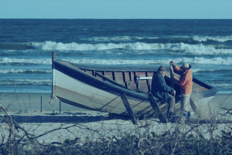 A imagem mostra um grupo de pescadores na praia, próximos a um barco de pesca. O barco está na areia e os pescadores estão conversando ao lado. Ao fundo, vê-se o mar com ondas e um céu claro. A vegetação rasteira está presente na área da praia.