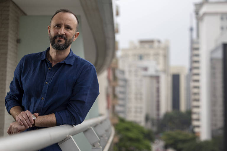 Um homem com barba e cabelo curto, vestindo uma camisa azul, está apoiado em uma varanda. Ao fundo, há uma vista de prédios altos e vegetação, indicando um ambiente urbano. O céu está nublado.