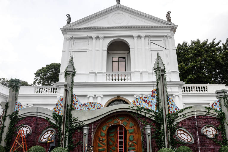 A imagem mostra a fachada de um edifício histórico, predominantemente branco, com detalhes arquitetônicos clássicos. A entrada principal é em forma de arco, com portas de madeira. Ao redor, há elementos decorativos coloridos e vegetação bem cuidada. No primeiro plano, uma escada de madeira está posicionada ao lado da entrada, e há grandes arbustos esféricos na frente.