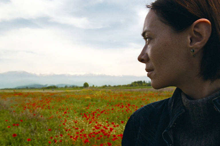 A imagem mostra uma mulher de perfil, observando um vasto campo coberto de flores vermelhas, possivelmente papoulas. Ao fundo, há uma cadeia de montanhas com neve visível e um céu nublado. A mulher tem cabelo curto e usa um suéter escuro, com um olhar contemplativo voltado para a paisagem.