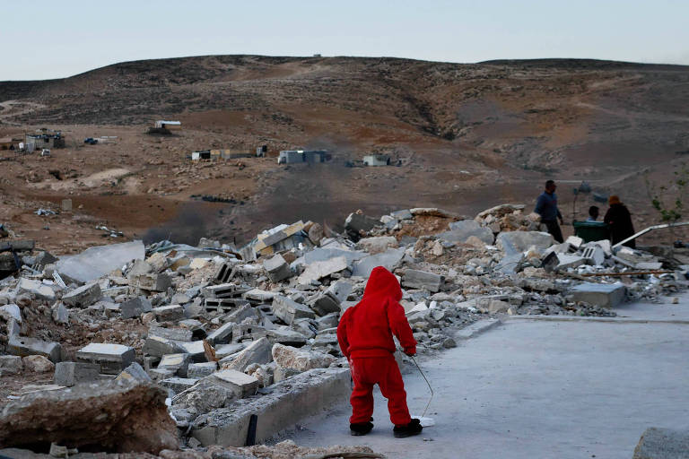 A imagem mostra uma criança vestindo um agasalho vermelho, de costas, caminhando sobre escombros em uma área devastada. Ao fundo, há colinas áridas e algumas construções danificadas. No lado direito, adultos estão em pé, observando a cena. O ambiente é desolado, com muitos destroços visíveis.