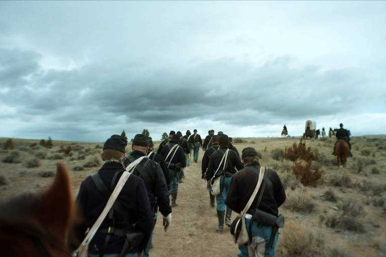 A imagem mostra um grupo de soldados marchando em uma paisagem árida. Eles estão vestidos com uniformes escuros e carregam mochilas e equipamentos. O céu está nublado, e ao fundo, há algumas árvores e carroças puxadas por cavalos. A cena transmite uma sensação de movimento e determinação.