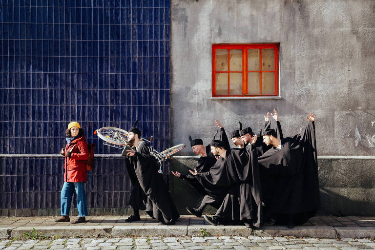 A imagem mostra uma cena de performance teatral ao ar livre. À esquerda, uma pessoa vestida com um casaco vermelho e um chapéu amarelo observa um grupo de seis pessoas vestidas com roupas pretas, que estão em poses dramáticas. Ao fundo, há uma parede cinza com uma janela vermelha e azulejos azuis. O ambiente é urbano e parece ser uma rua.