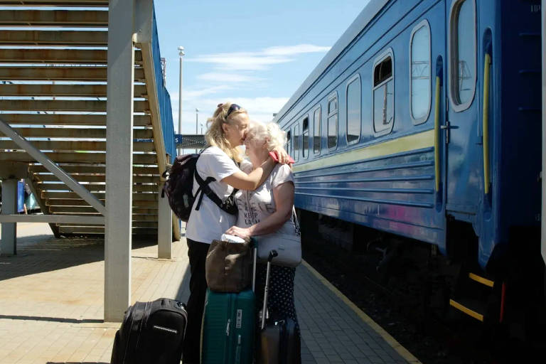 A imagem mostra duas mulheres se abraçando e se beijando em uma estação de trem. Uma delas é mais jovem, com cabelo loiro e usa uma camiseta branca e calças escuras, enquanto a outra é mais velha, com cabelo grisalho e usa uma camiseta clara e calças com estampa. Ambas estão próximas a malas de viagem. Ao fundo, há um trem azul e uma escada de metal. Outras pessoas estão visíveis na estação.