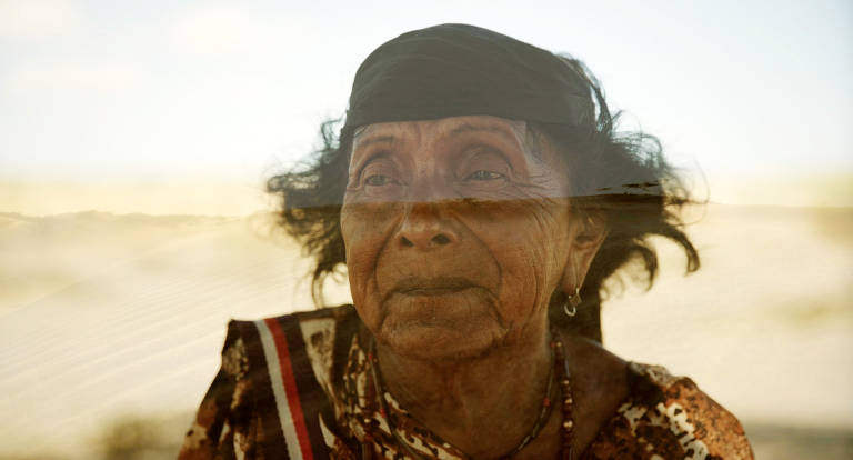 A imagem mostra o rosto de uma mulher idosa com cabelo encaracolado e uma faixa preta na cabeça. Ela está vestindo uma blusa colorida e parece estar em um ambiente ao ar livre, com um fundo desfocado que sugere um cenário natural. A expressão dela é serena e contemplativa.