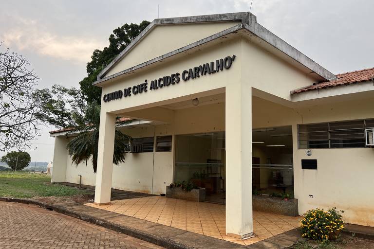 A imagem mostra a fachada do Centro de Café Alcides Carvalho, um edifício de um andar com uma entrada ampla. O nome do centro está escrito em letras grandes e pretas na parte superior da entrada. O piso é de cerâmica em tons de bege e há um pequeno jardim com flores amarelas na frente. Ao fundo, pode-se ver árvores e uma área verde.