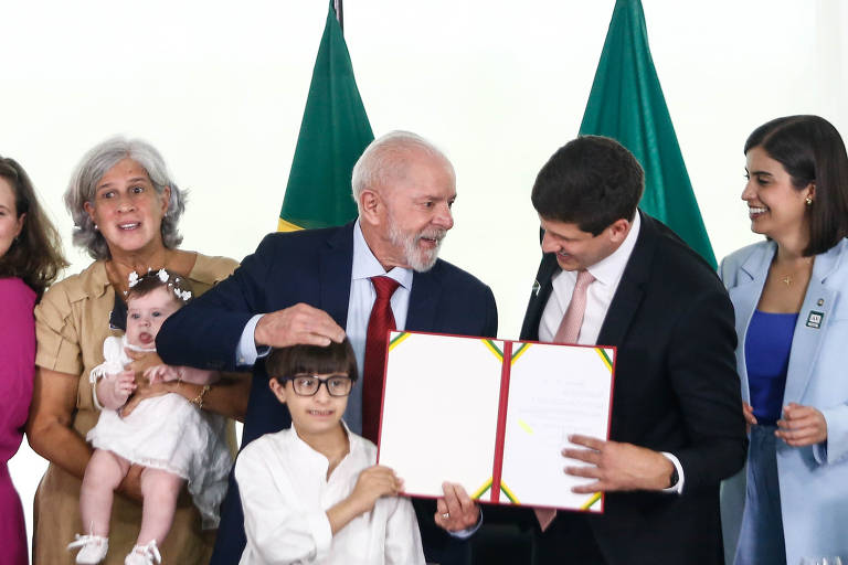 A imagem mostra um grupo de pessoas em uma cerimônia de assinatura. No centro， um homem de cabelo grisalho e barba， vestido com um terno escuro e gravata vermelha， está sorrindo e segurando um documento. Ao seu lado， um homem mais jovem， também de terno， segura o documento. Uma criança de camisa branca está ao lado deles， enquanto uma mulher com um bebê no colo e outras pessoas observam ao fundo. Bandeiras do Brasil estão visíveis ao fundo.
