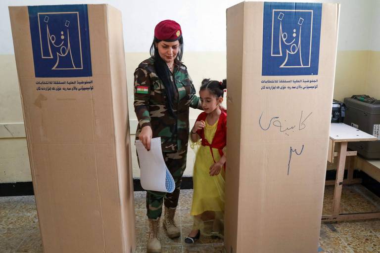 A imagem mostra uma mulher vestida com uniforme militar e um boné vermelho, ao lado de uma menina que usa um vestido amarelo. Elas estão em um local de votação, com cabines de votação ao fundo, que possuem símbolos eleitorais. A mulher segura um papel e parece estar ajudando a menina.