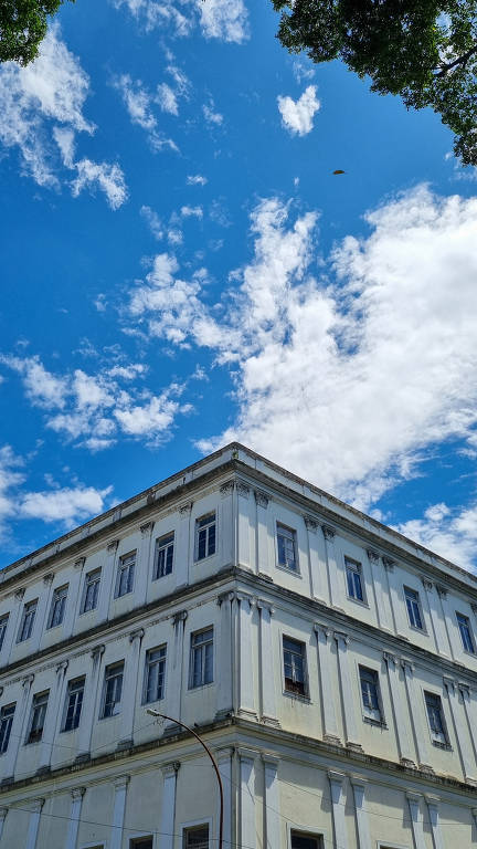 A imagem mostra a parte superior de um edifício de cor clara, com janelas e detalhes arquitetônicos visíveis. O céu ao fundo é azul, com nuvens brancas dispersas. A perspectiva é de baixo para cima, destacando a estrutura do edifício contra o céu.