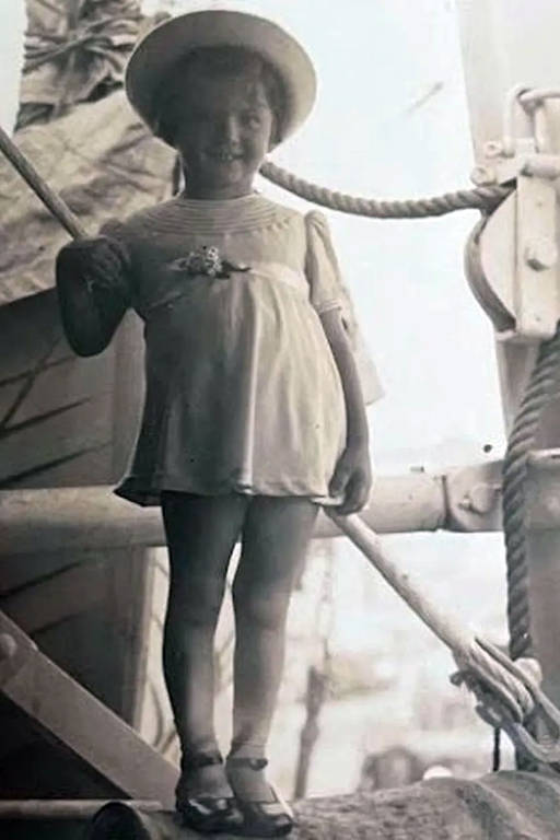 A imagem mostra uma menina sorridente em pé sobre uma estrutura de madeira em um barco. Ela usa um vestido claro e um chapéu de palha. A menina segura uma vara com uma mão e está descalça, com os pés visíveis. Ao fundo, há elementos de um barco, como cordas e partes de madeira