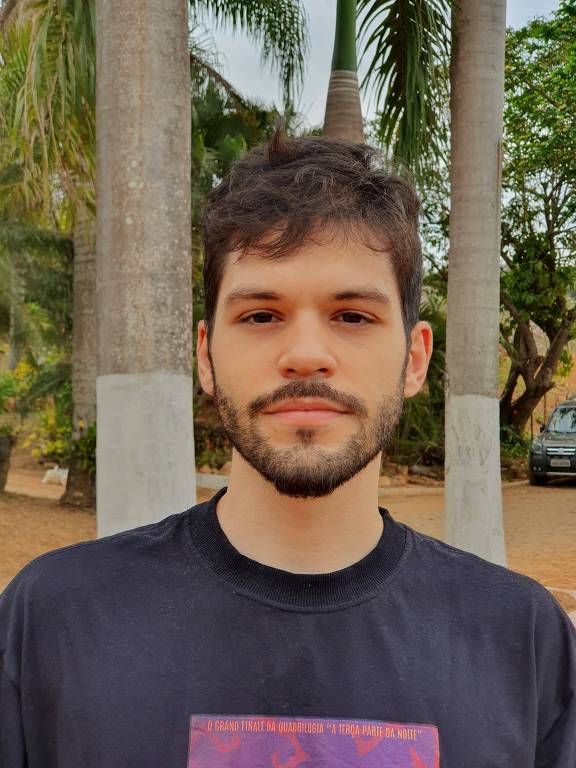 Retrato de Arthur, um homem branco com cabelo castanho claro e barba curtos está posando para a foto em um ambiente ao ar livre. Ao fundo, há palmeiras. Ele usa uma camiseta escura com um design em tons de roxo na parte frontal.