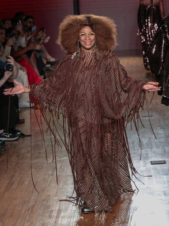 ma modelo caminha na passarela durante um desfile de moda, vestindo um traje marrom com franjas. Ela tem cabelo volumoso e está sorrindo, com os braços abertos. Ao fundo, há uma plateia de pessoas assistindo, algumas segurando câmeras. Outras modelos podem ser vistas ao fundo, usando roupas brilhantes.